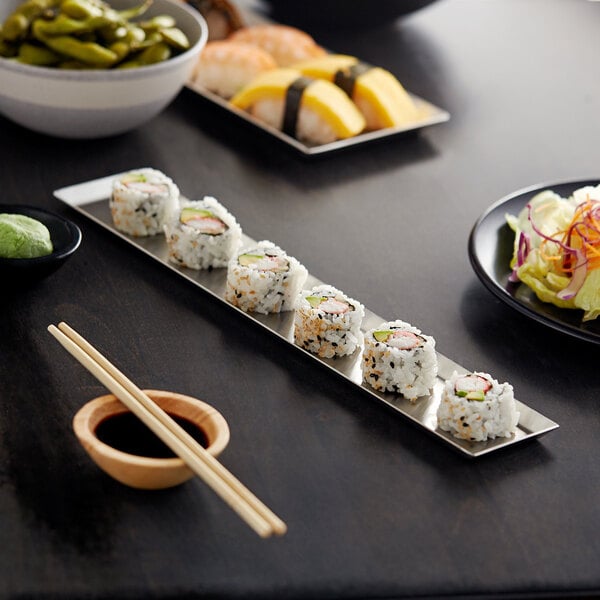 A rectangular stainless steel tray with sushi and a wooden bowl of sauce on a table.
