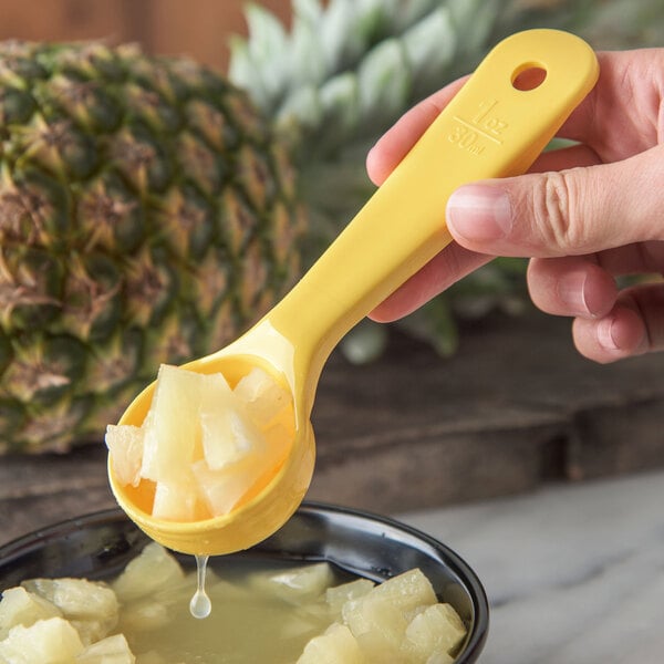 A person holding a Carlisle yellow perforated portion spoon filled with pineapple chunks and juice.