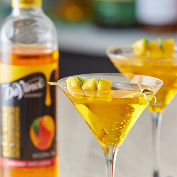 Two martini glasses filled with mango flavored drinks on a table with a bottle of DaVinci Gourmet Mango Syrup.