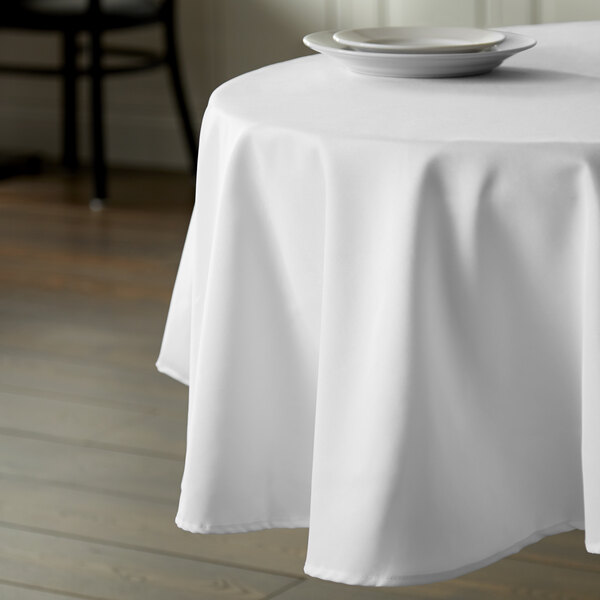 A table with a white Intedge 100% polyester tablecloth and white plates on it.