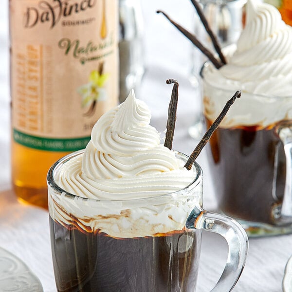 A glass mug of hot chocolate with whipped cream and a vanilla stick.