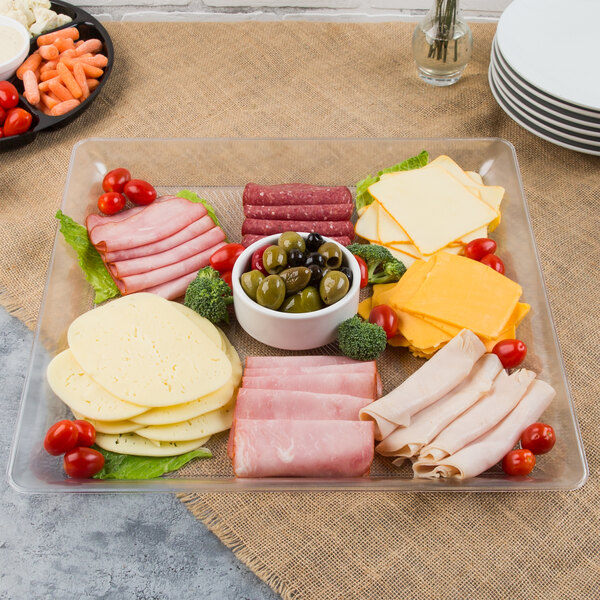 A Fineline clear plastic square catering tray with meat, cheese, olives and vegetables on a table.