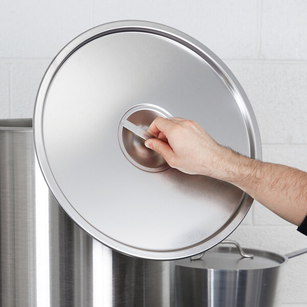 A hand holding a Vollrath stainless steel lid over a metal pot.