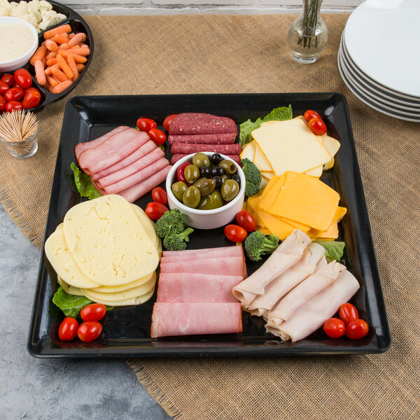 A Fineline black plastic square cater tray with meat, cheese, and vegetables on a table.