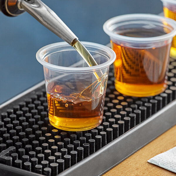 A person pouring brown liquid from a pipette into clear plastic shot glasses on a table.