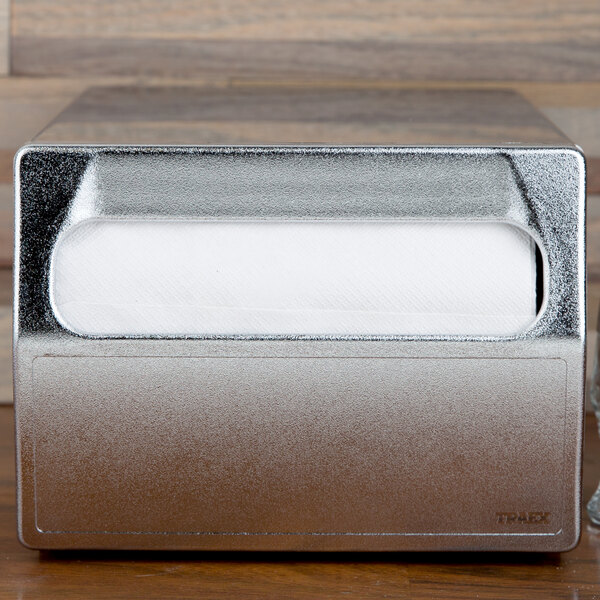 A Vollrath stainless steel countertop napkin dispenser with a white napkin in it.