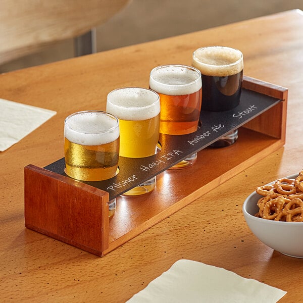 An Acopa wooden tasting flight tray with four pub glasses of beer and a bowl of pretzels on a table.