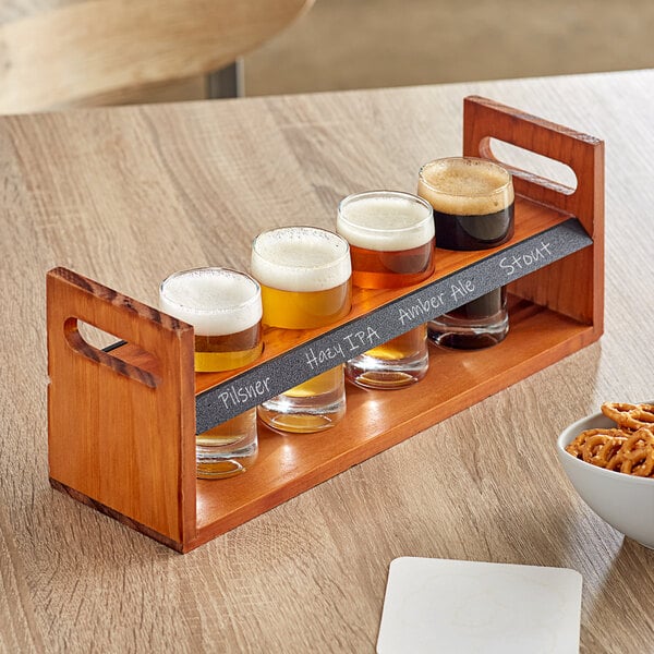 An Acopa wooden flight carrier with four pub tasting glasses of beer on a wood table with a bowl of pretzels.