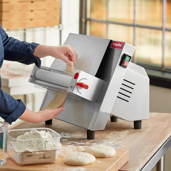 A woman using an Estella countertop dough sheeter to roll pizza dough on a counter.
