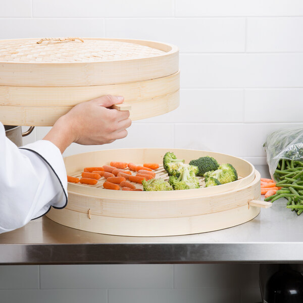 A person using a Town bamboo steamer to cook a bowl of baby carrots, broccoli, and other vegetables.