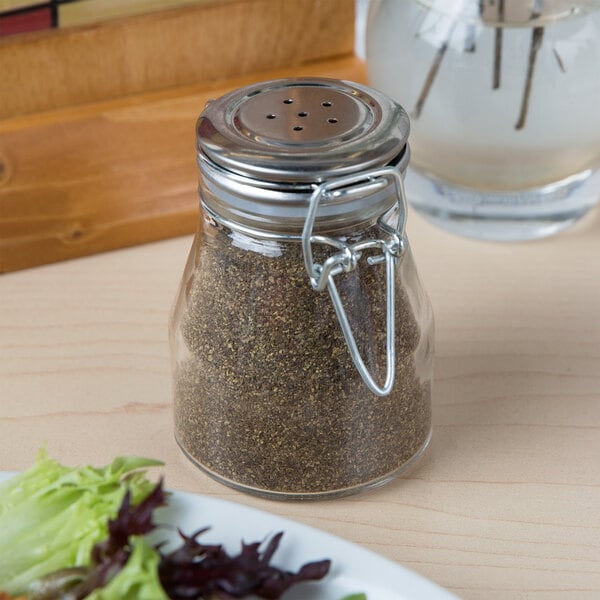 A Tablecraft glass salt and pepper shaker jar with a metal clip-top lid on a table with a plate of salad.