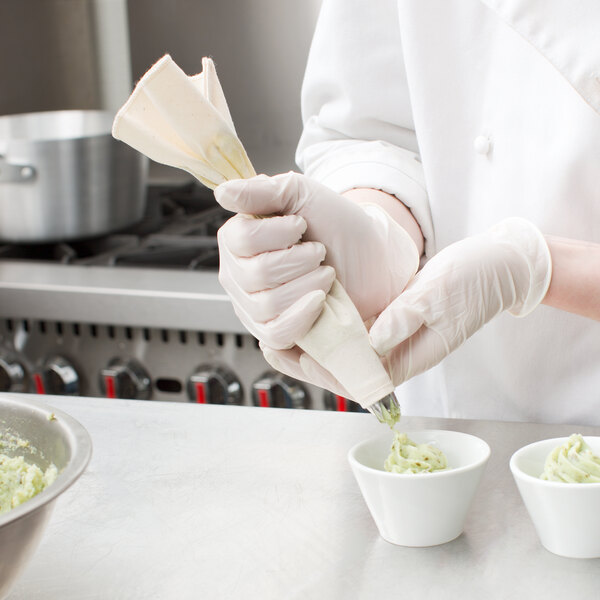 A person wearing white gloves using an Ateco canvas pastry bag to pipe green food into a white bowl.
