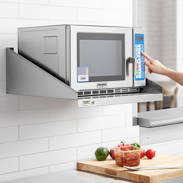 A person pointing at a microwave on a Regency stainless steel wall mount shelf.