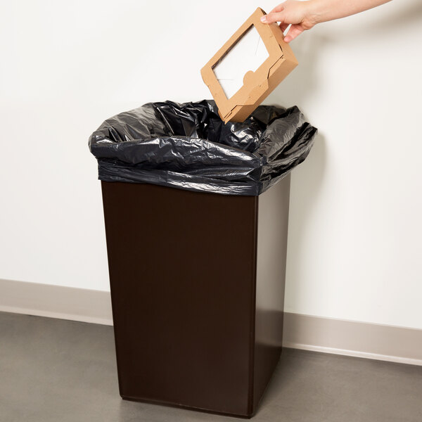 A hand placing a cardboard box in a brown Continental Swingline trash can with a black plastic bag inside.