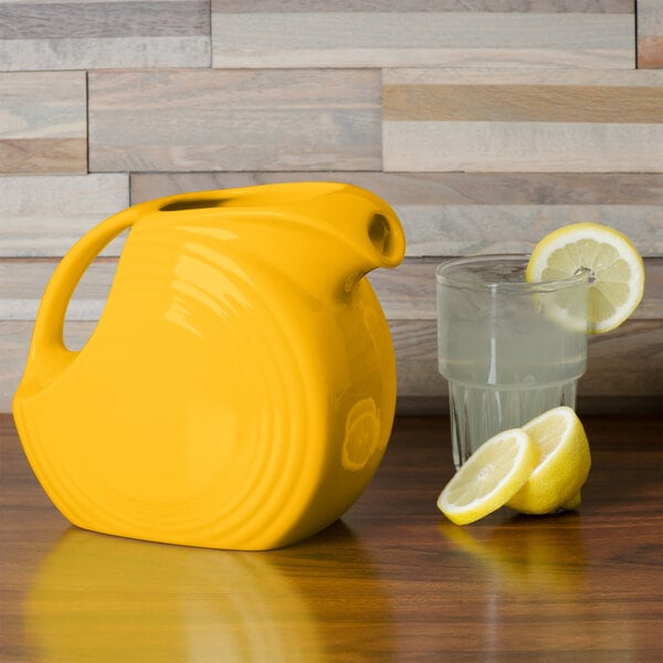 A yellow Fiesta Disc China pitcher on a counter with a glass of water and lemons.