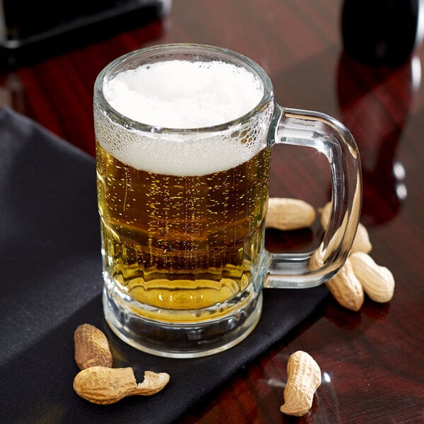 A Libbey beer mug filled with beer and peanuts on a table.