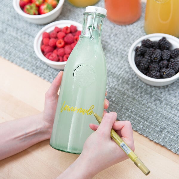A person using a yellow Franmara mini tip marker to write on a glass bottle of orange juice.