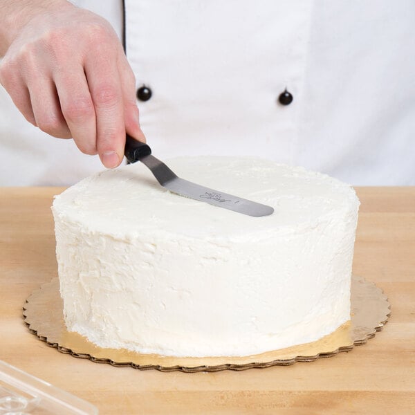 A hand using an Ateco offset spatula with a black handle to cut a cake.