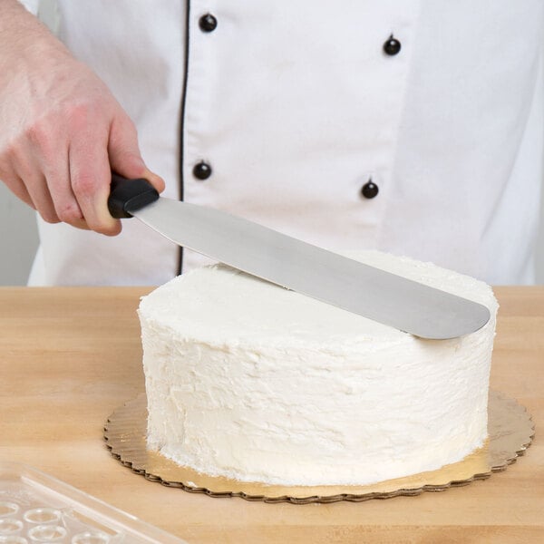 A hand holding a black Ateco baking spatula cutting a cake.