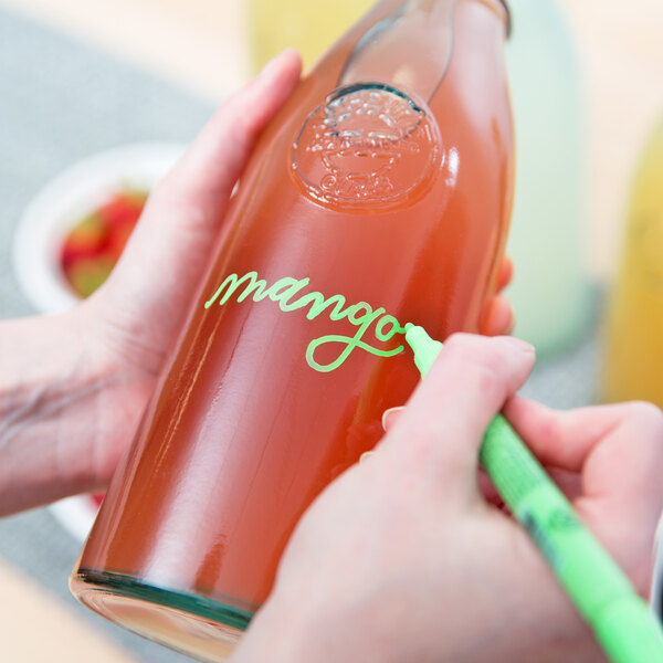 A hand using a Franmara neon green mini tip glass marker to write on a bottle of liquid.