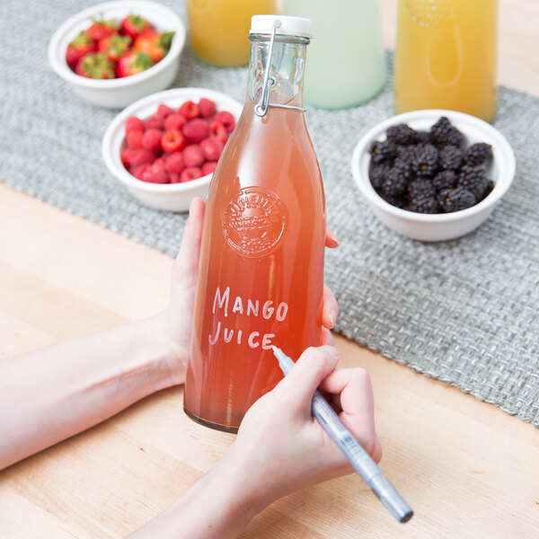 A person using a Franmara neon silver mini tip glass marker to write on a bottle of juice.