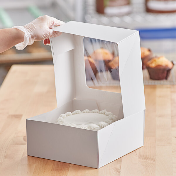 A gloved hand holding a white window cake box with a cake inside.