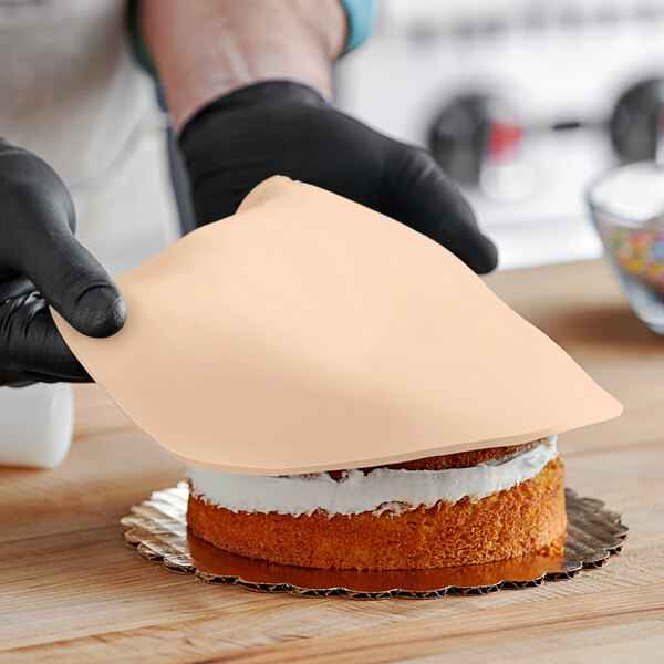 A person in black gloves using a tan object to cut a cake with peach fondant on it.