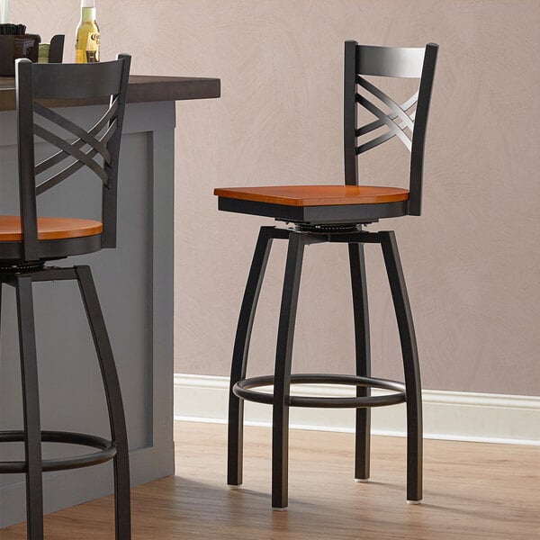 A pair of Lancaster Table & Seating black cross back swivel bar stools with cherry wood seats.