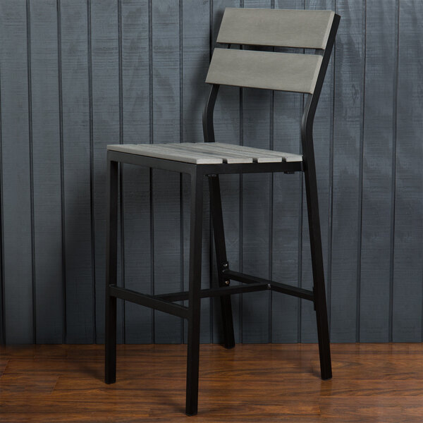 A BFM Seating black aluminum outdoor restaurant bar stool with gray synthetic teak back and seat against a wall.