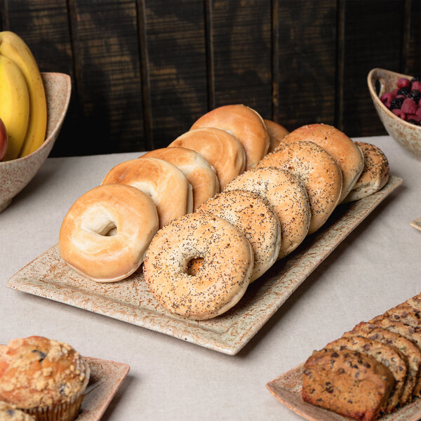 A rectangular porcelain platter with bagels, muffins, and fruit on display.