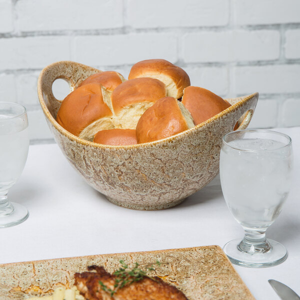 A white porcelain bowl with cut-out handles filled with bread on a table with plates of food.