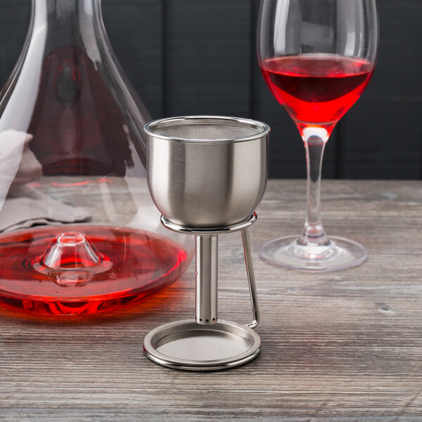 A Franmara stainless steel decanter funnel on a table stand next to a glass of red liquid.