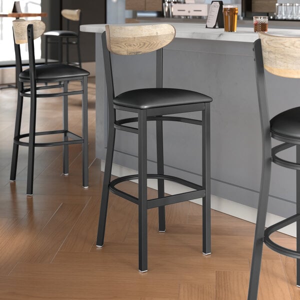 Lancaster Table & Seating Boomerang Series Black Finish Bar Stool with Black Vinyl Seat and Driftwood Back at a restaurant counter.