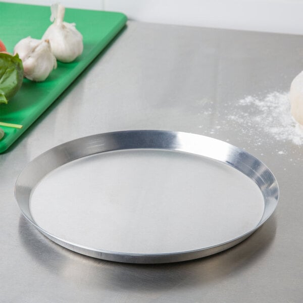 An American Metalcraft heavy weight aluminum pizza pan with a white surface next to vegetables.