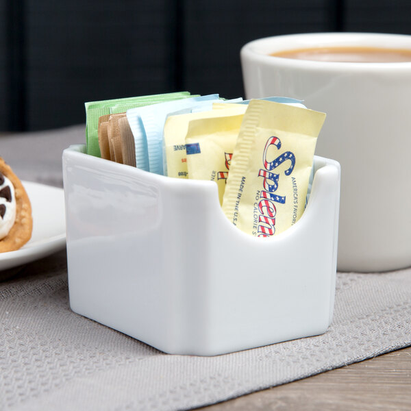 A white Libbey porcelain sugar caddy on a counter with sugar and coffee packets inside.