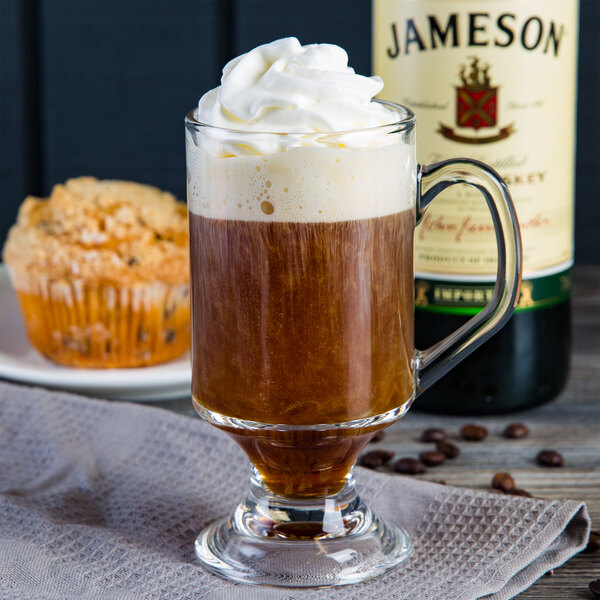 A fully tempered Arcoroc Irish coffee mug filled with brown liquid and whipped cream on a table with a muffin.