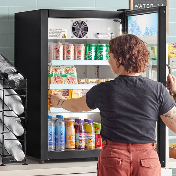 A woman opening an Avantco black countertop display refrigerator with a swing door.