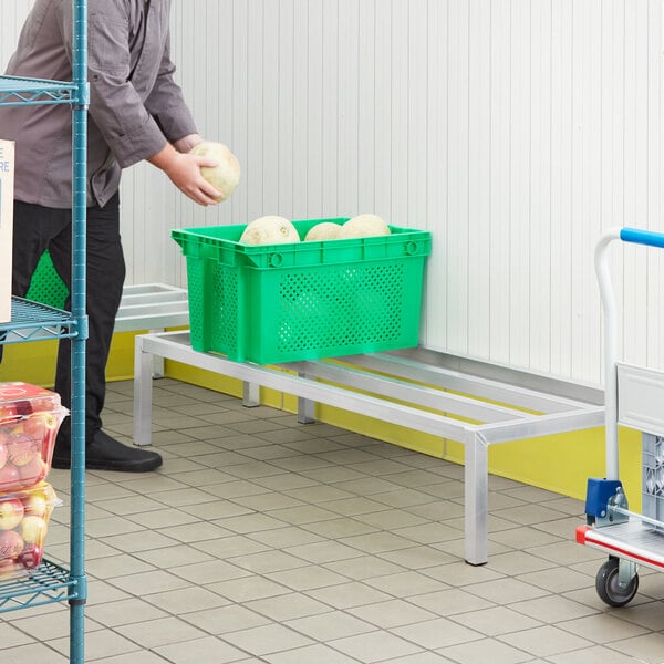 A man putting a green basket of melons on a Regency aluminum dunnage rack in a room with a cart and boxes.