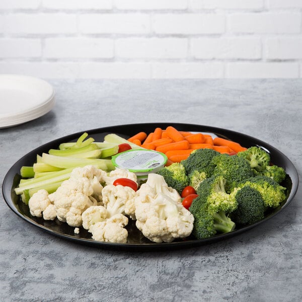 A WNA Comet black round catering tray with broccoli, carrots, and tomatoes on it.