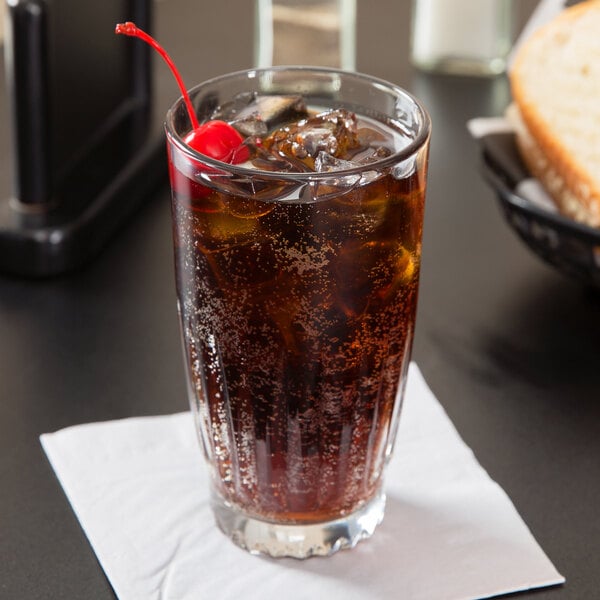 A Libbey Winchester beverage glass filled with soda, ice, and a cherry on a table in a bar.