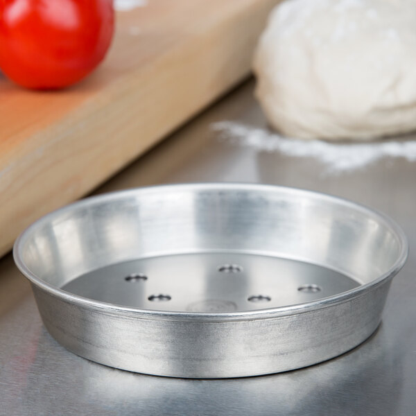 An American Metalcraft tin-plated steel pizza pan with dough and tomatoes on it.