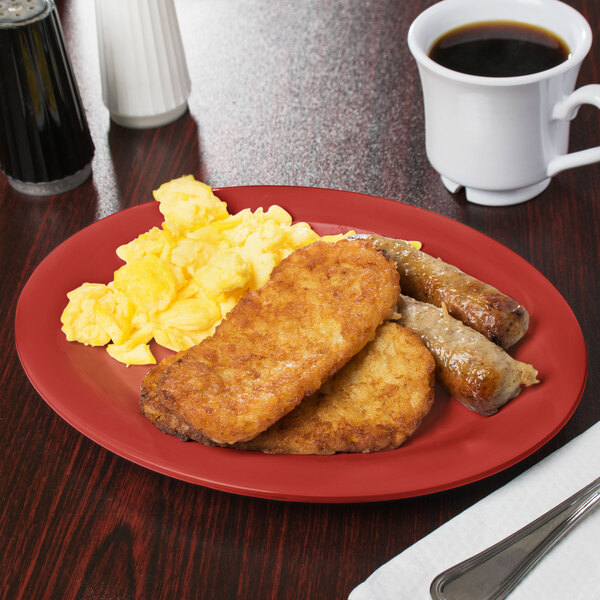 A cranberry oval platter with food including scrambled eggs, sausage, and toast on a table with a cup of coffee.
