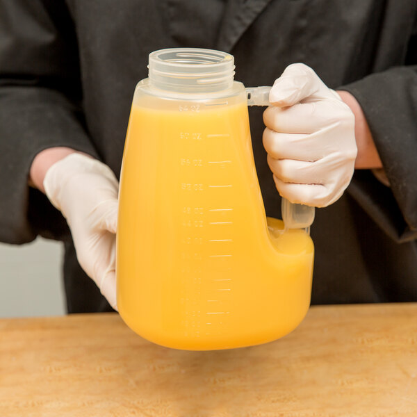 A person in gloves holding a Tablecraft 64 oz. dispenser jar filled with yellow liquid.