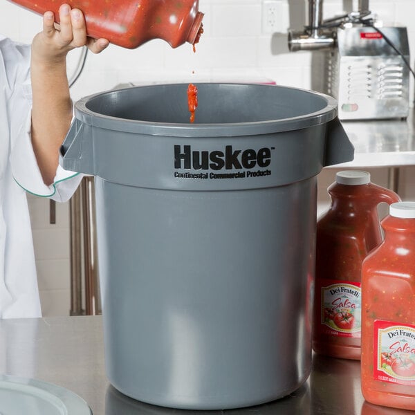 A woman in a white coat pouring red sauce into a Continental 10 gallon gray round trash can.