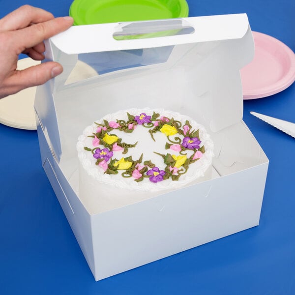 A hand holding a white bakery box with a cake decorated with flowers inside.