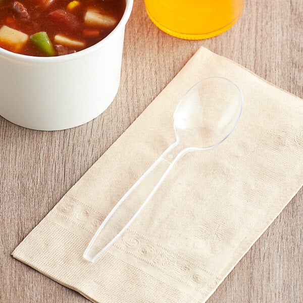 A Visions clear heavy weight plastic soup spoon on a napkin next to a bowl of soup.
