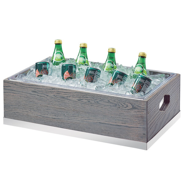 A Cal-Mil gray oak wood ice housing with bottles of soda in ice.