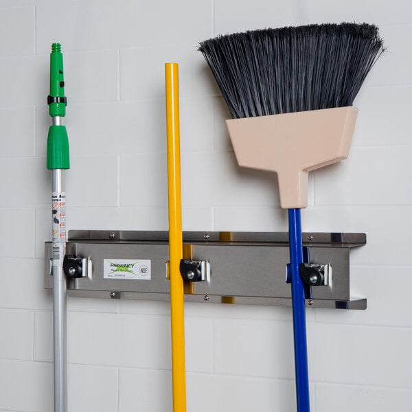 A Regency stainless steel mop and broom rack with 3 holders on a wall with a broom and mop hanging from it.