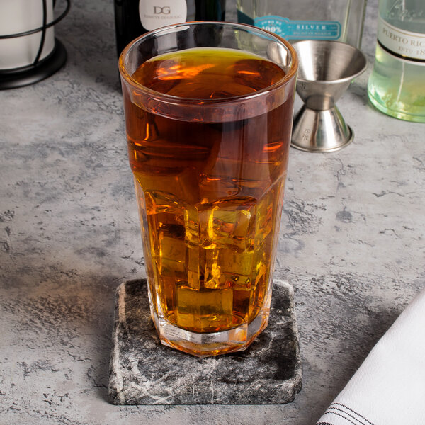 An Arcoroc Gotham cooler glass of amber liquid with ice cubes on a stone coaster.