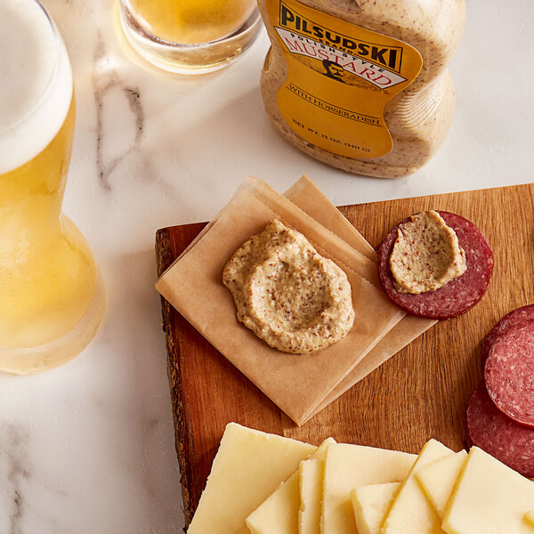 A cutting board with cheese, sausage, and Pilsudski Polish Style Horseradish Mustard next to a glass of beer.